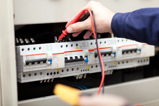 Intérieur professionnel, technicien en veste bleue effectuant mesures électriques sur tableau de distribution